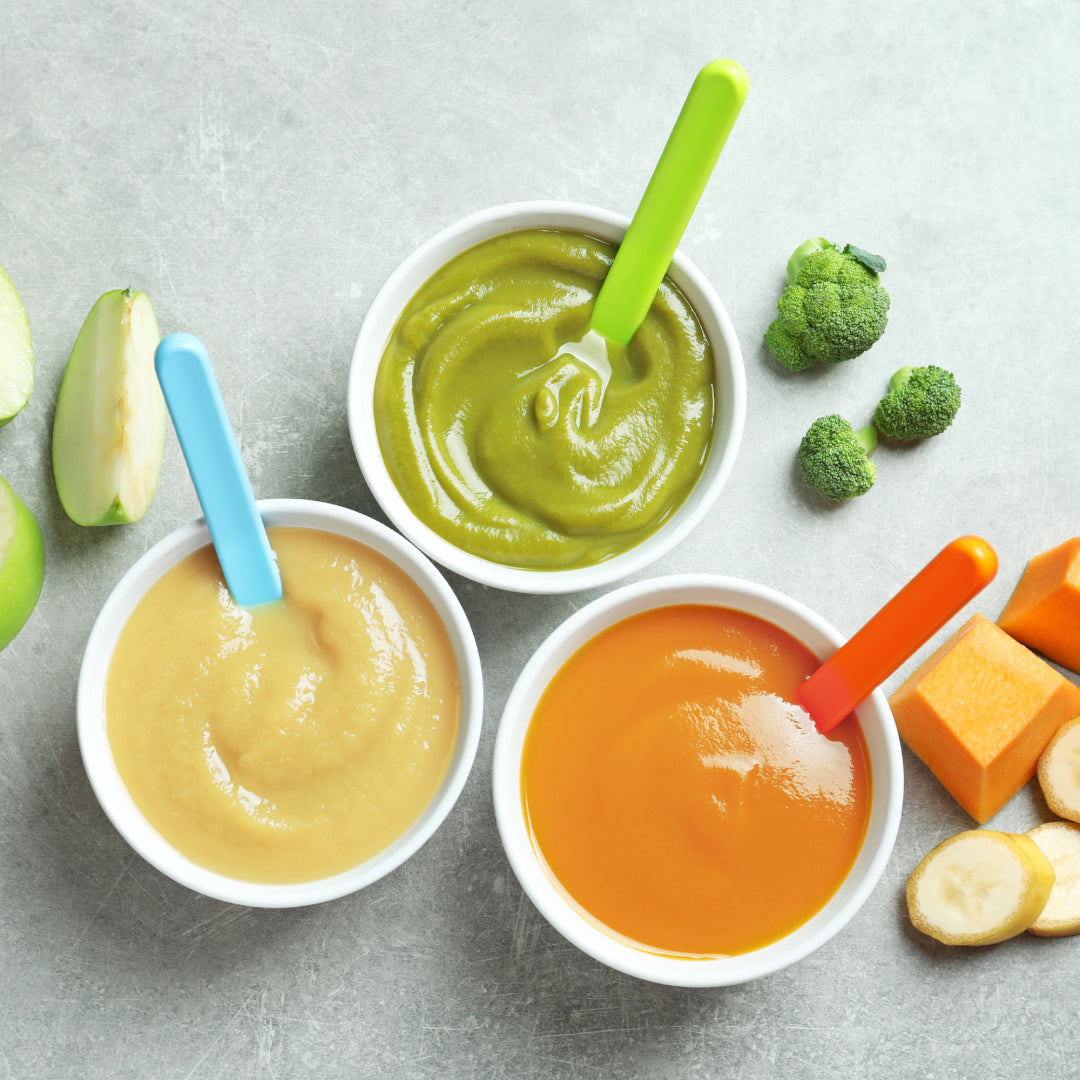 Three bowls of baby food -  puréed apple, one puréed broccoli, one puréed mango and banana. Fresh ingredients are next to the bowls