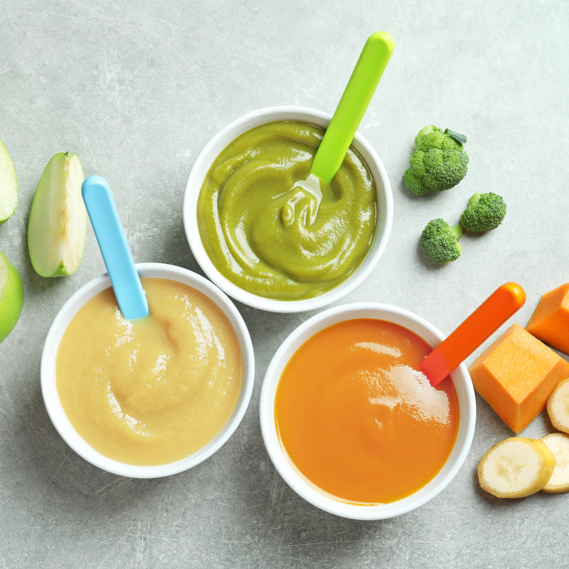 Three bowls of baby food -  puréed apple, one puréed broccoli, one puréed mango and banana. Fresh ingredients are next to the bowls