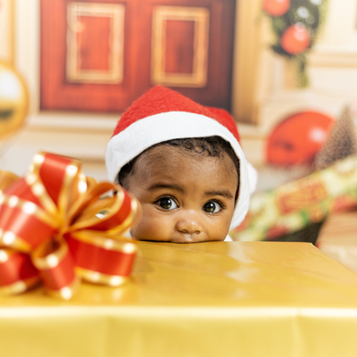 A cute baby wearing a Santa hat is biting on the edge of a gold paper-wrapped present