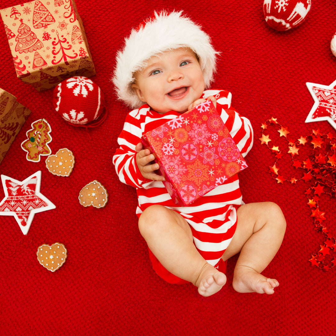 A smiling baby lying on their back, dressed in red & white clothes and a Santa hat. The baby is holding a present wrapped in red.