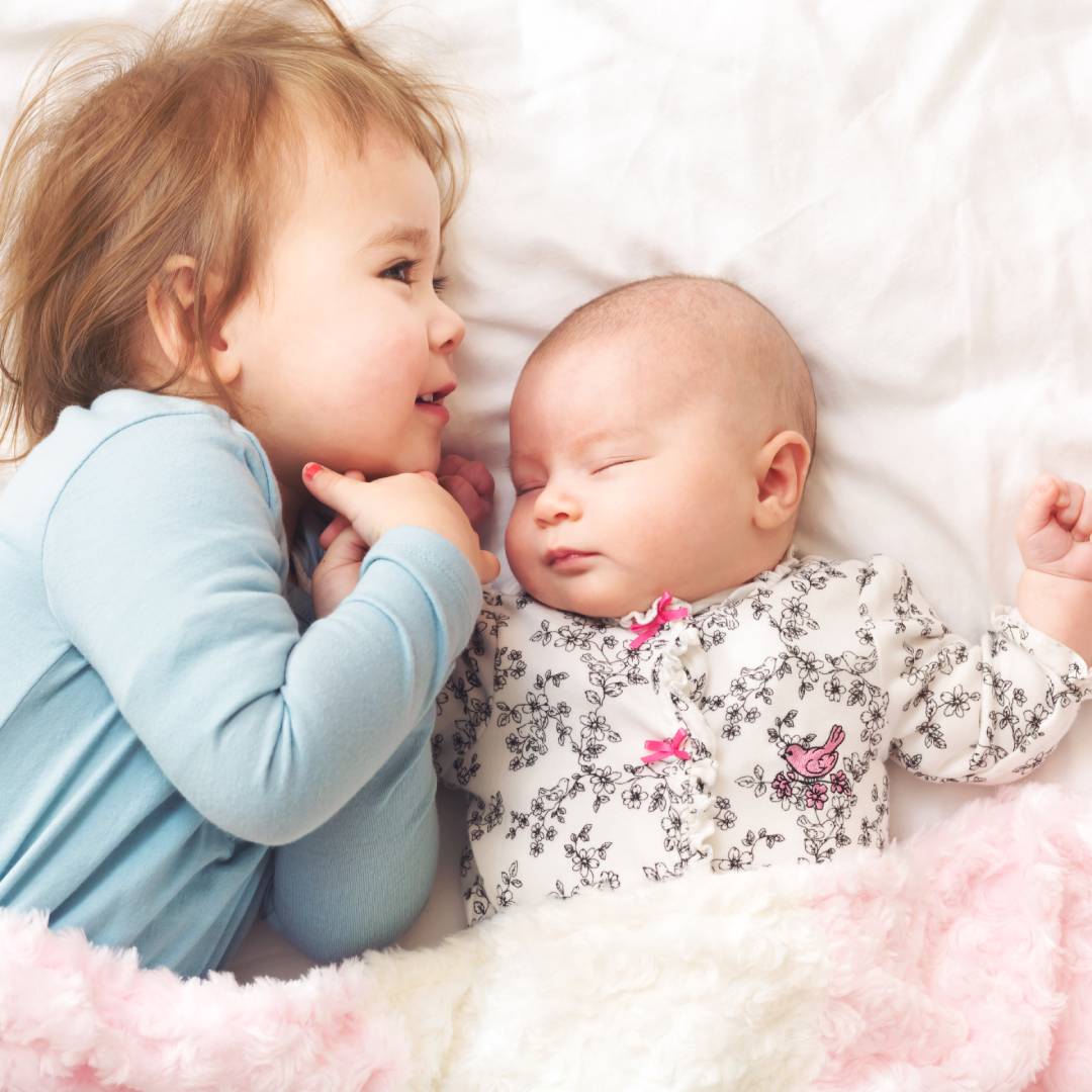 Smiling toddler holding the hand of their sleeping newborn sibling